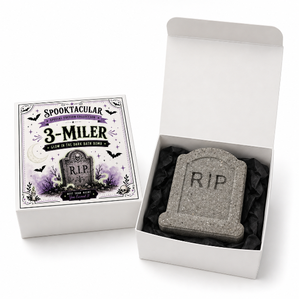 A Halloween-themed bath bomb set shown in its packaging, featuring a gray tombstone-shaped bath bomb with 'RIP' engraved inside an open white box, alongside the illustrated product box labeled 'SPOOKTACULAR 3-MILER'.